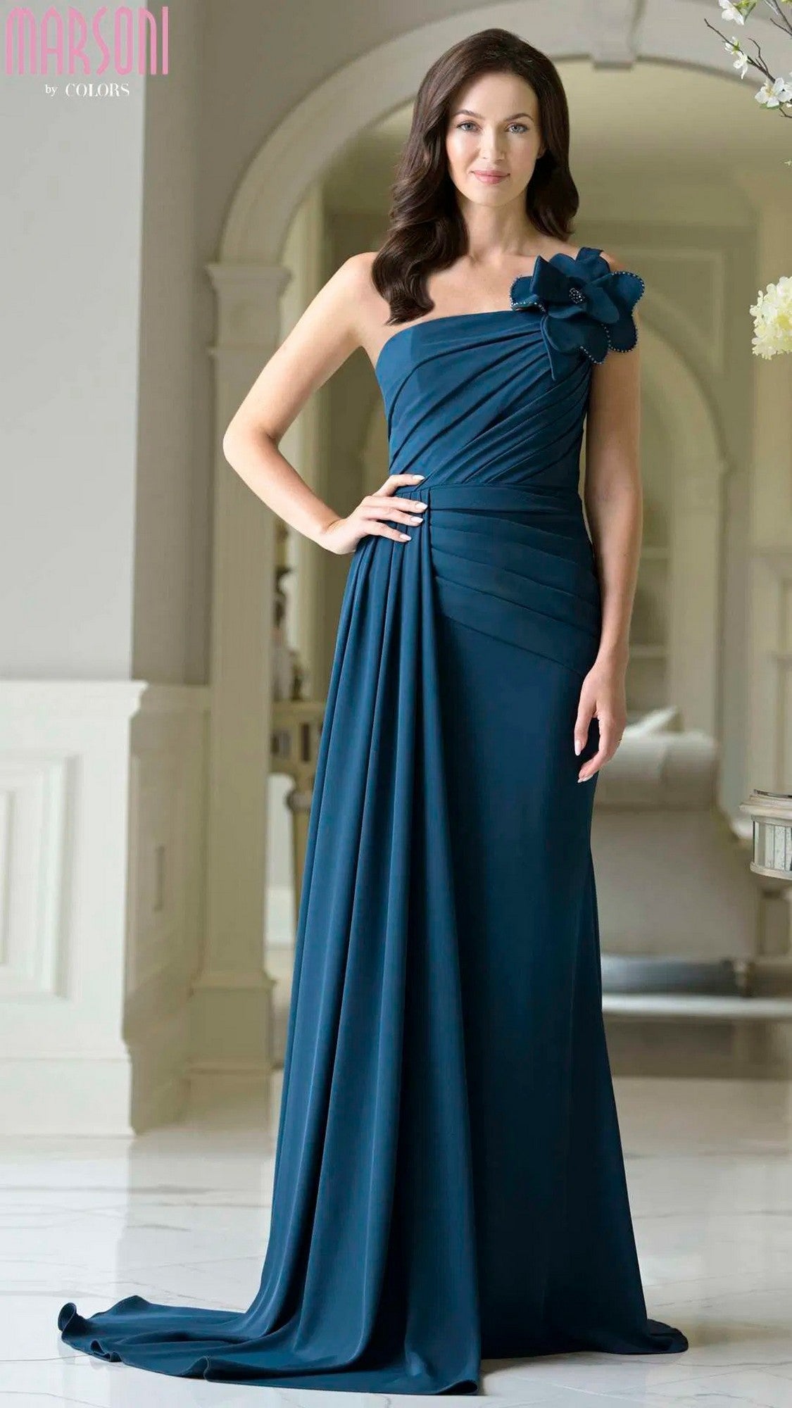 A woman stands indoors in a Marsoni M330 long formal dress, one-shoulder style in peacock color, highlighted by a striking 3D floral accent on the shoulder.